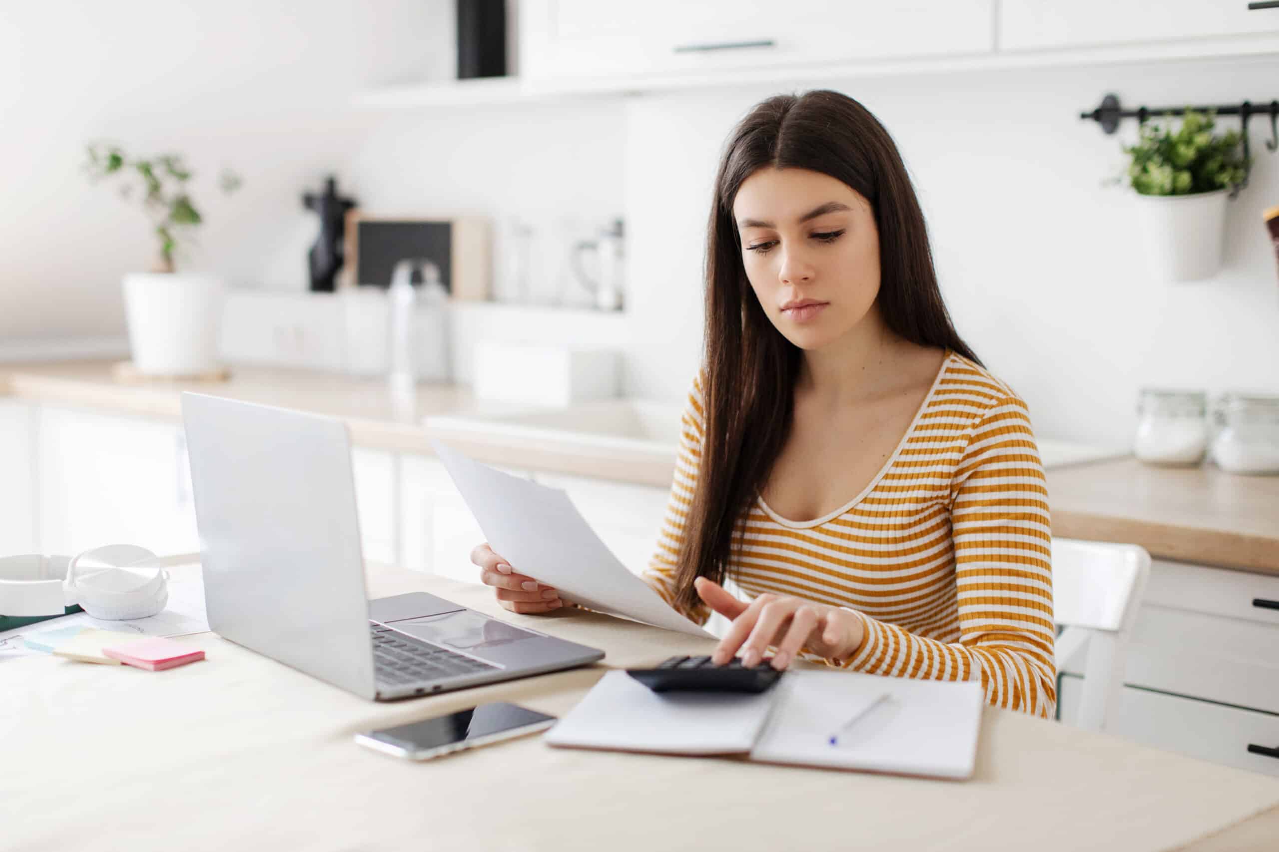 Une femme, assise à la table de la cuisine, remplit des documents administratifs à l'aide d'une calculatrice, calcule les impôts et établit un budget. Elle vérifie des documents financiers.