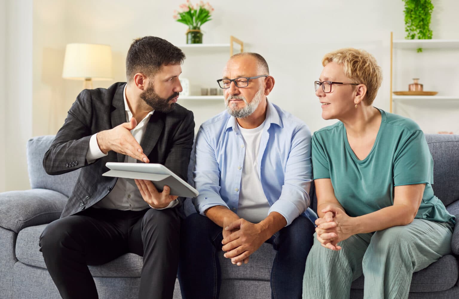 Un agent immobilier passe en revue les détails d'un contrat sur sa tablette tandis qu'un couple d'âge mûr l'écoute depuis son canapé lors d'une visite de logement. Un conseiller masculin donne des conseils à des personnes âgées sur les placements et les assurances.