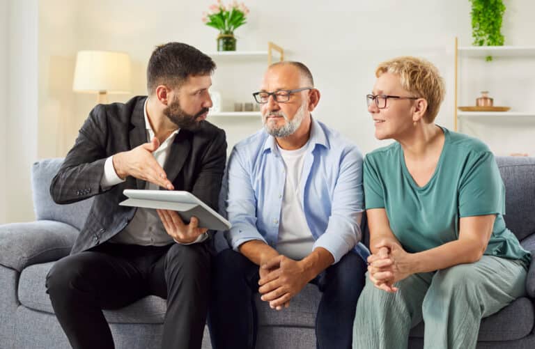 Un agent immobilier passe en revue les détails d'un contrat sur sa tablette tandis qu'un couple d'âge mûr l'écoute depuis son canapé lors d'une visite de logement. Un conseiller masculin donne des conseils à des personnes âgées sur les placements et les assurances.