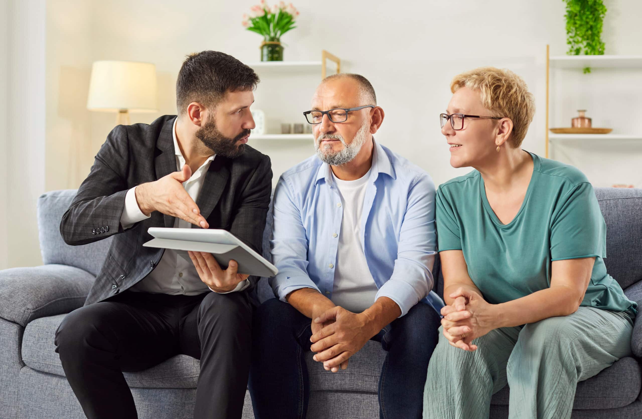 Un agent immobilier passe en revue les détails d'un contrat sur sa tablette tandis qu'un couple d'âge mûr l'écoute depuis son canapé lors d'une visite de logement. Un conseiller masculin donne des conseils à des personnes âgées sur les placements et les assurances.
