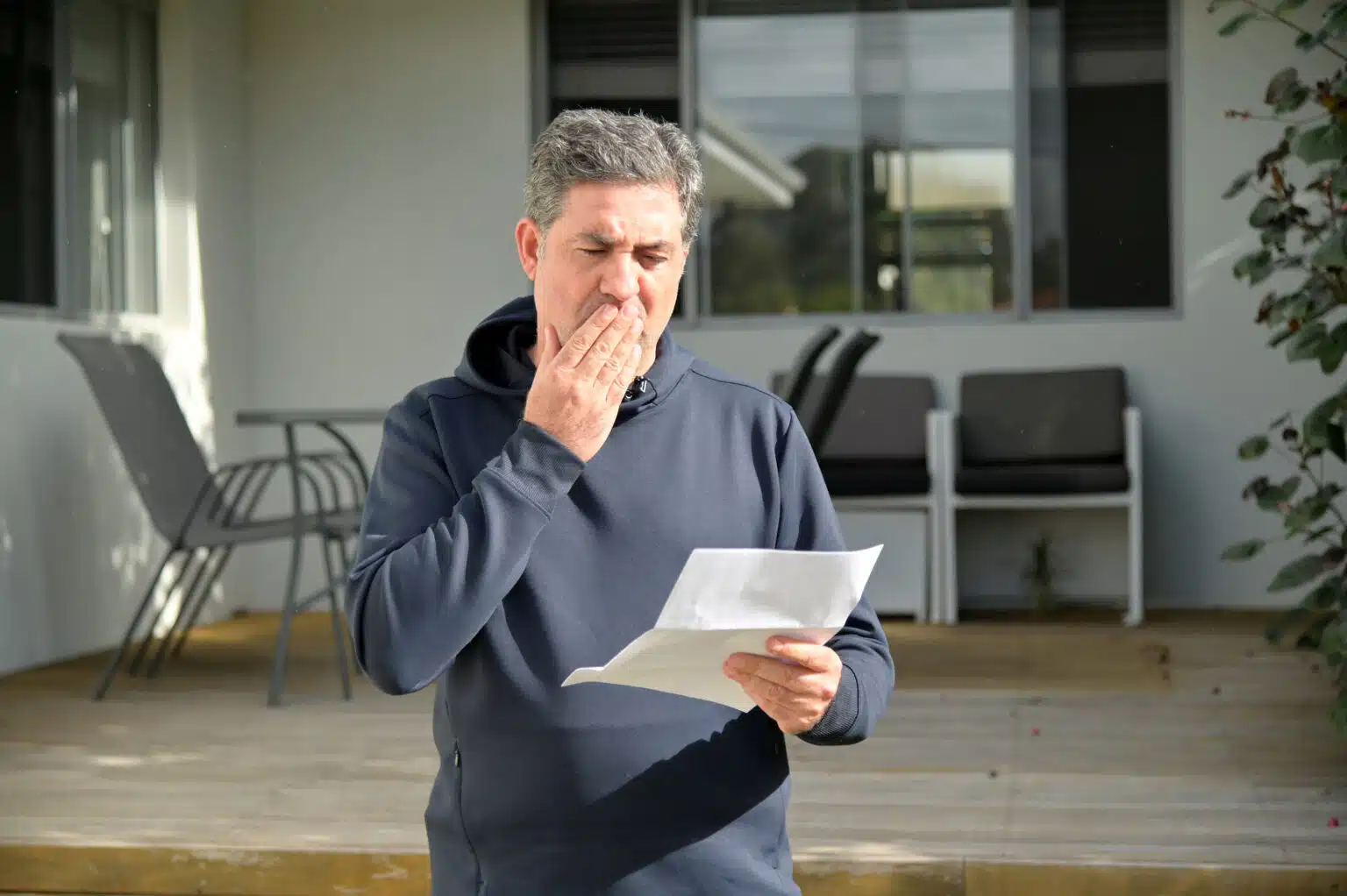 Un homme d'âge mûr (entre 40 et 50 ans), l'air choqué, en train de lire une lettre devant la porte d'entrée d'une maison.